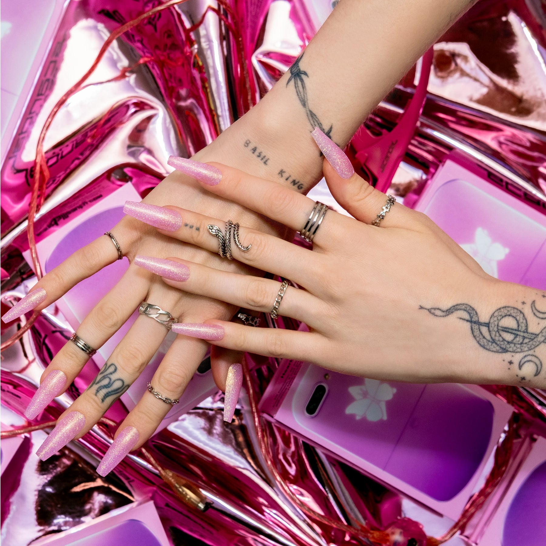 Two hands overlapping wearing baby pink press-on nails with fine iridescent holographic glitter above holographic pink 2000's era inspired background.