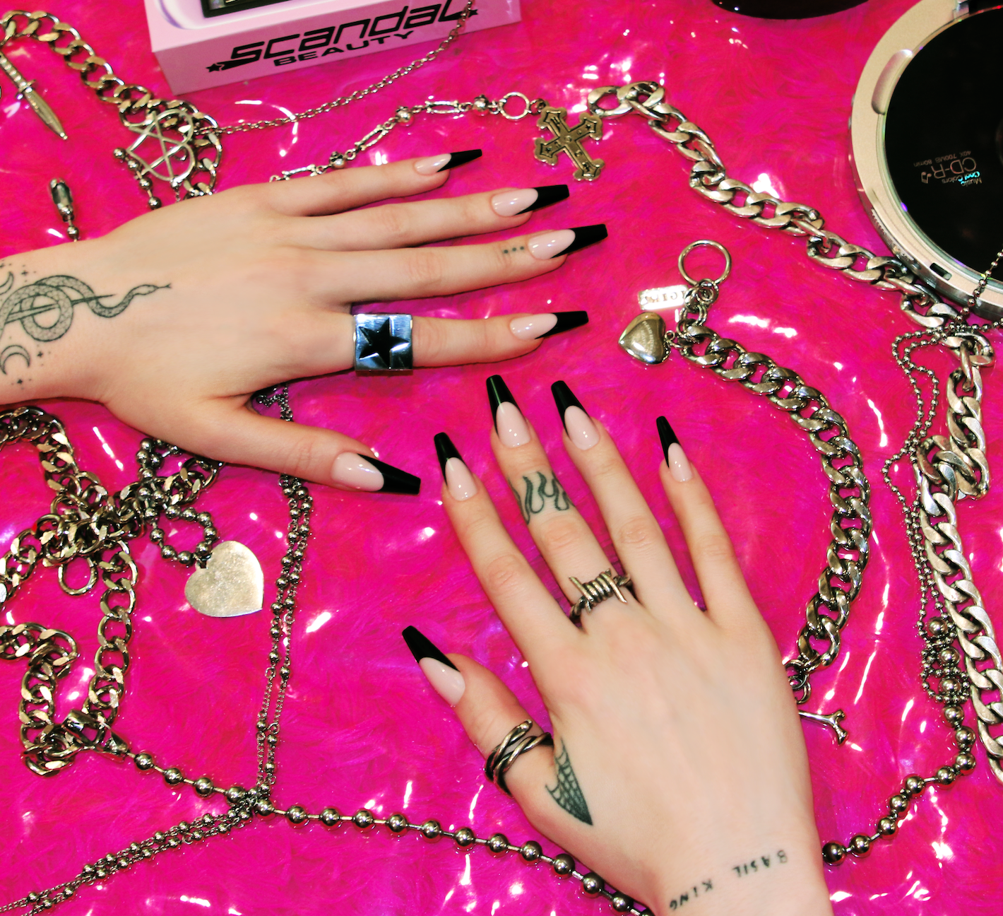 Two hands wearing black French tip nails surrounded by 2000's alternative era inspired silver metal jewelry chains, featuring a CD player. On hot pink fur background.